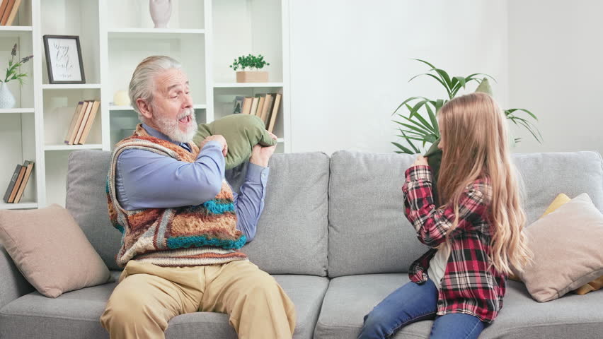 Carefree grandfather and child resting on cozy couch and playfully throwing pillows at each other. Cheerful man of mature age fooling around with restless kid in modern living room during weekend. Royalty-Free Stock Footage #1104296701