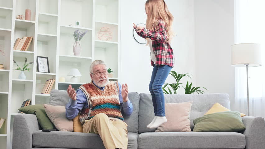Restless long-haired blonde girl jumping on grey sofa in fashionable spacious living room. Tired grandfather complaining on migraine and touching head with hands. Royalty-Free Stock Footage #1104296731