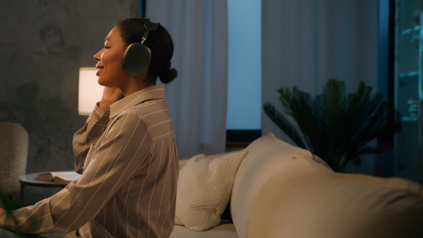 Tired African American woman female relax on sofa in evening living room enjoy audio with wireless headphones lean on comfortable couch rest eyes closed leisure listen relaxing music sing song smile Royalty-Free Stock Footage #1104298027