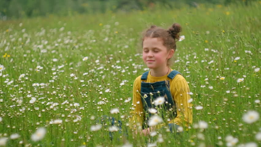 Happy adorable preteen girl kid sitting on grass opens eyes and enjoy amazing warm nature at summer day. Dreamy peaceful relaxed smiling child breathing fresh air on beautiful big chamomile fields