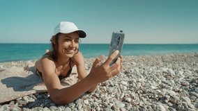 relaxed woman tourist in bikini sunbathes on sea beach close to water, unlocks her mobile phone and takes photo or video call. Blue Lagoon resort, Oludeniz, Turkey.