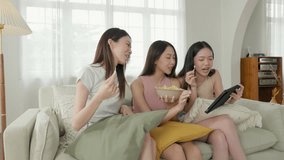 Group of young adult Asian females, with beautiful healthy skin, enjoying potato chips and a tablet content, laughing and having fun together at home. showcasing their strong bond of friendship.