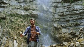 Happy male tourist in trip, journey. Young man blogger traveler takes selfie photo or video for social media of amazing waterfall using phone. 4k slow motion footage. Wanderlust, travel concept.