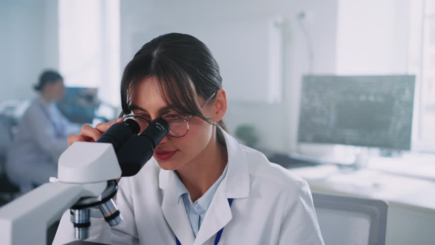 Close-up of young brunette woman wearing scientist uniform looking at camera. Attractive girl using modern microscope and experimenting in bright laboratory room. Daytime, working day. Job concept Royalty-Free Stock Footage #1104329085