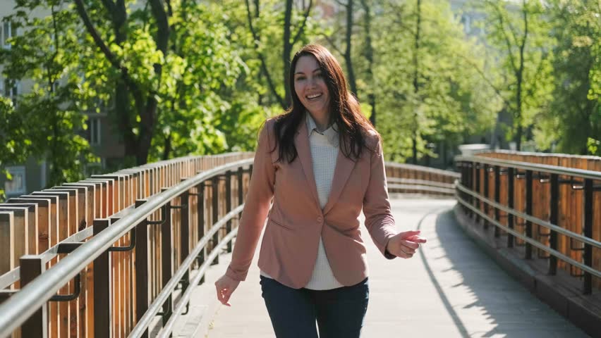 A beautiful brunette woman in a pink suit smiles, has fun and dances on the street during the day.
