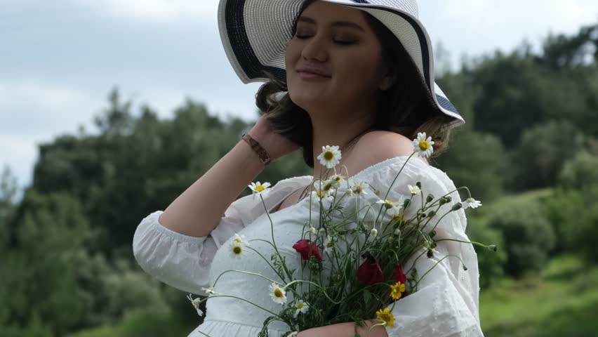 A close-up of a pregnant woman in a white dress and hat, putting a daisy into her ear while holding freshly picked daisies and poppies in a wheat field