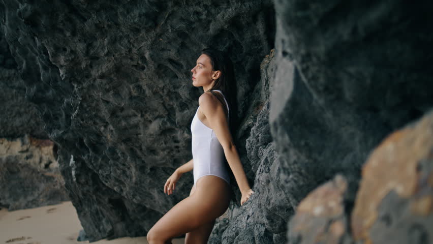 Hot young woman posing on rocky coast leaning at stone cliff. Attractive sensual model looking camera confidently wearing white sexy swimsuit. Beautiful serious girl standing on seashore cloudy day.