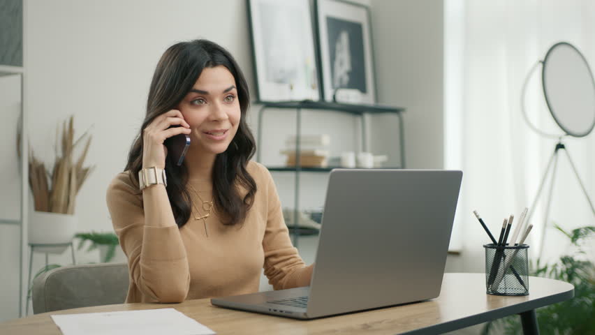 Smiling young hispanic business woman talking on phone using laptop sits at home office desk. Happy latin female customer make mobile call confirming online website shopping order delivery concept 4K Royalty-Free Stock Footage #1104339845