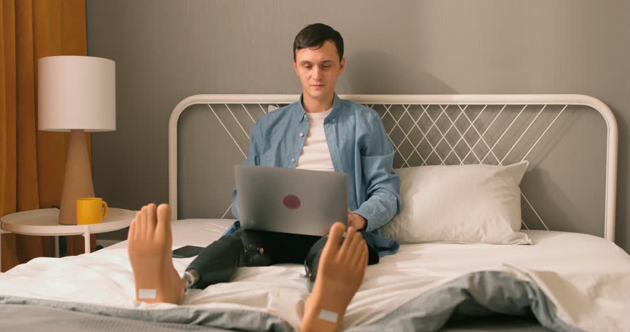 Young man with prosthesis working online, having lesson drinking tea, having break, rest, sitting on bed. Confident caucasian guy with physical disability having online work. never give up concept | Shutterstock HD Video #1104344165