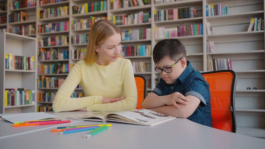 Slow motion. A school boy with Down's syndrome, sitting at the desk with his tutor in the library campus, learning, reading encyclopedia. Education and social integration of children with disabilities Royalty-Free Stock Footage #1104344545