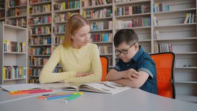 Slow motion. A school boy with Down's syndrome, sitting at the desk with his tutor in the library campus, learning, reading encyclopedia. Education and social integration of children with disabilities - Powered by Shutterstock - Get 15% off with code: PIKWIZARD15