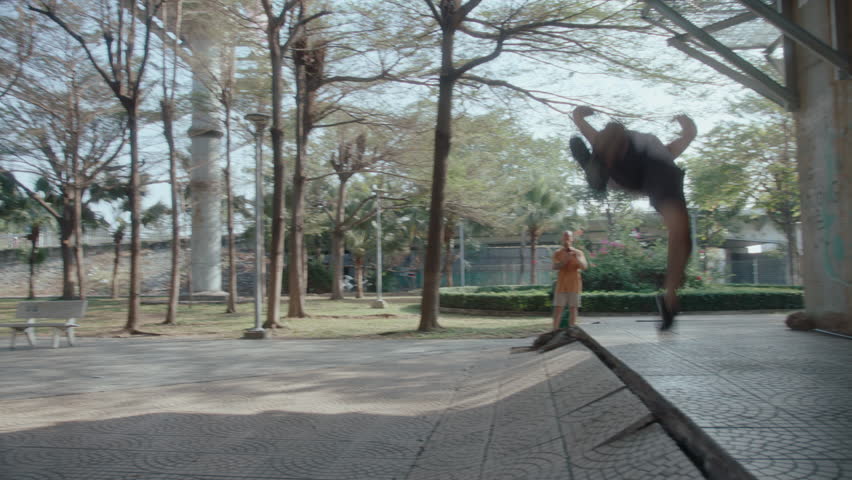 Parkour athlete performing backflip stunt, practicing outdoors in city park on summer day, his friend standing behind and filming video with phone. Slow motion wide shot - Powered by Shutterstock - Get 15% off with code: PIKWIZARD15