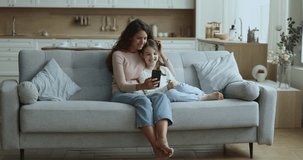 Beautiful Latina woman hugs her little cute daughter showing her funny mobile app, make selfie photos, watch videos on-line content, laughing rest together on sofa at home with modern cellphone gadget