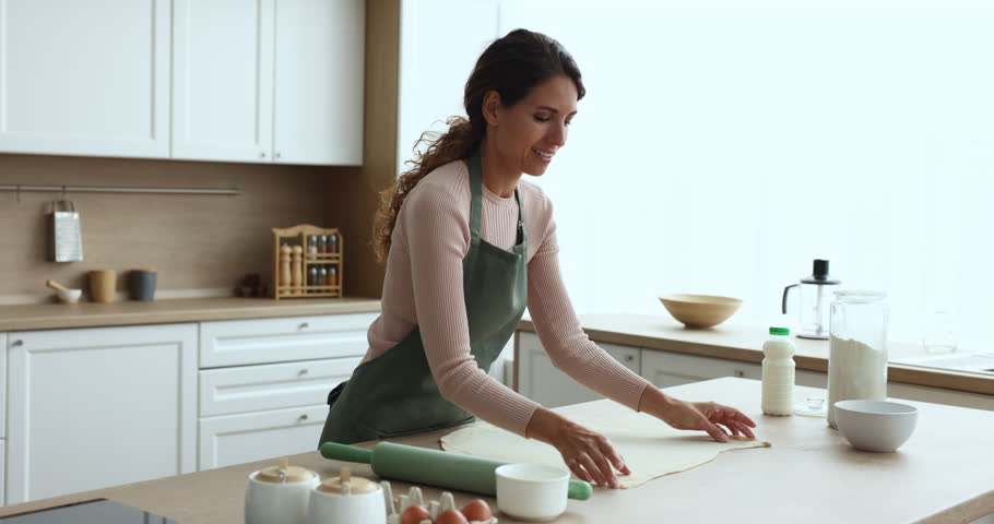 Smiling woman housewife in apron spend time in kitchen flattening dough, preparing bakery food, sweet dessert for family dinner, enjoy homemade cooking, culinary hobby. Routine and household chores Royalty-Free Stock Footage #1104349055