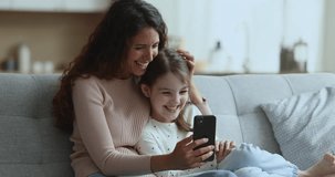 Young happy woman and little daughter laugh on funny videos, use new cool mobile application, enjoy videocall to relatives, spend pastime at home with modern app and smart phone. Family leisure, tech