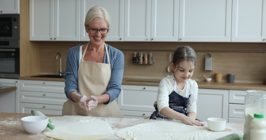 Happy mature grandmother and little granddaughter in aprons having fun, baking together, preparing dough clapping hands sprinkling white flour, laughing enjoy family time cooking holiday pie at home Royalty-Free Stock Footage #1104349103