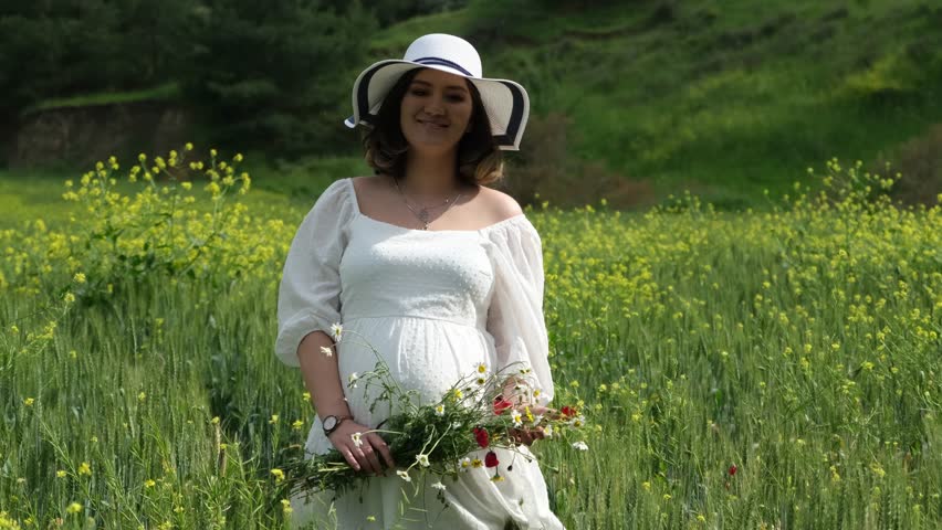 A close-up of a pregnant woman in a white dress and hat and holding freshly picked daisies and poppies in a wheat field