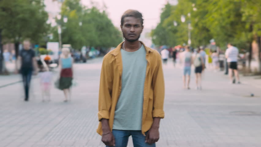 Time lapse portrait of Indian guy standing in busy street with stream of people moving around. Busy life and young people concept.