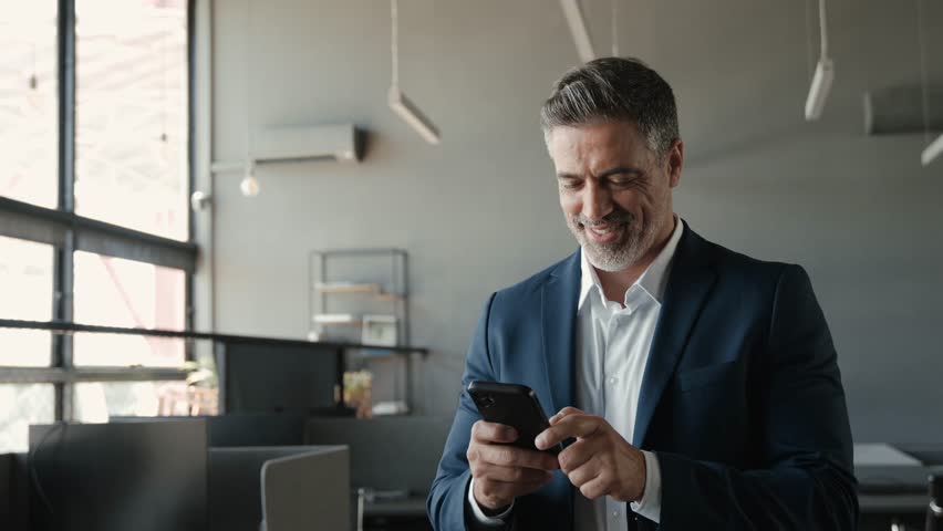 Smiling happy confident mid aged male company ceo executive wearing suit holding cellphone standing in office using business mobile apps technology financial online solutions on cell phone. | Shutterstock HD Video #1104365193