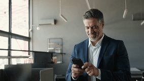 Smiling happy confident mid aged male company ceo executive wearing suit holding cellphone standing in office using business mobile apps technology financial online solutions on cell phone. - Powered by Shutterstock - Get 15% off with code: PIKWIZARD15
