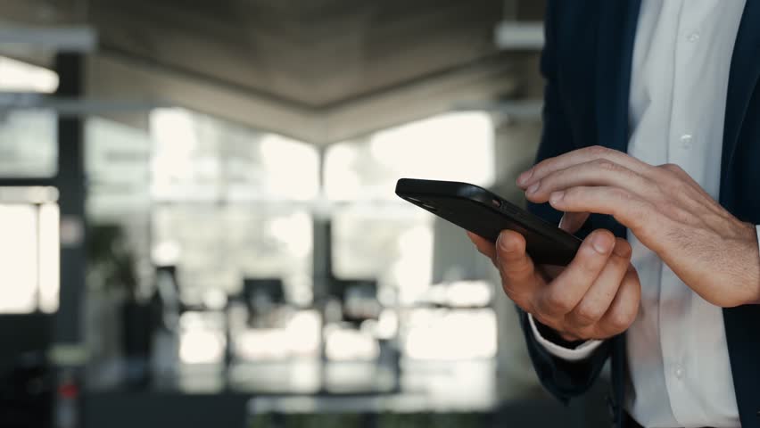 Smiling mid aged business man wearing suit standing in office using cell phone. Mature businessman professional executive holding smartphone in hand checking new mobile solution on cellphone. Close up Royalty-Free Stock Footage #1104365209
