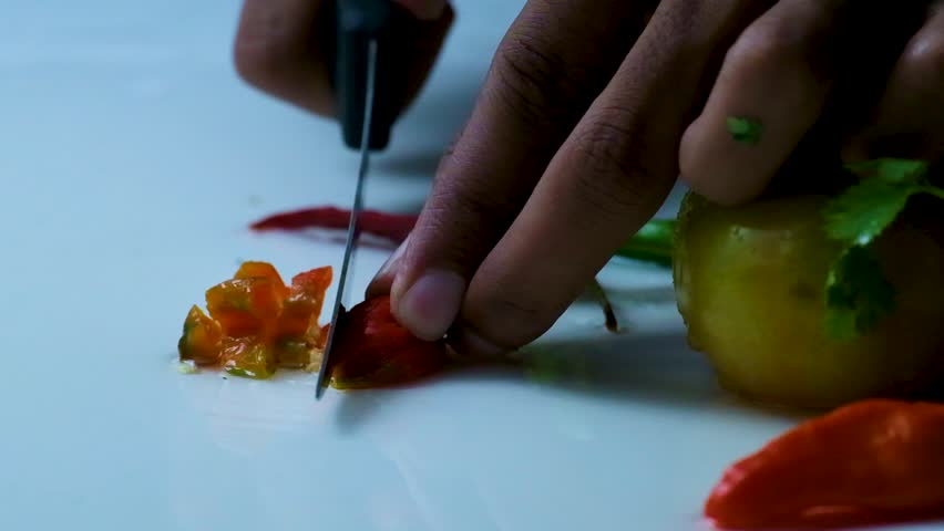 Slow Motion Shot of a Man Slicing a Red Bombay Chili Pepper on a White Surface next to Uncut Tomatoes