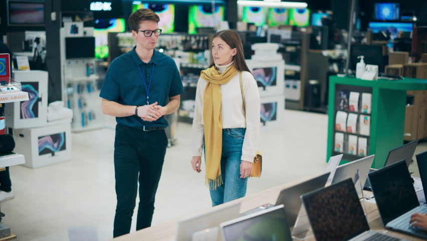 Young Woman Having a Conversation with a Salesperson in Home Electronics Store while Selecting a Laptop. Stylish Customer Wishes to Buy a Computer Device. Shopper Explores Latest Options with Expert Royalty-Free Stock Footage #1104370605