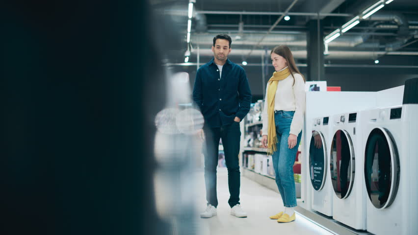 Stylish Young Man and Woman are Washing Machine Shopping at Home Retail Electronics Store. Couple Choosing a Laundry Cleaning Appliance for their Home. Customers Evaluate Options in Modern Shop