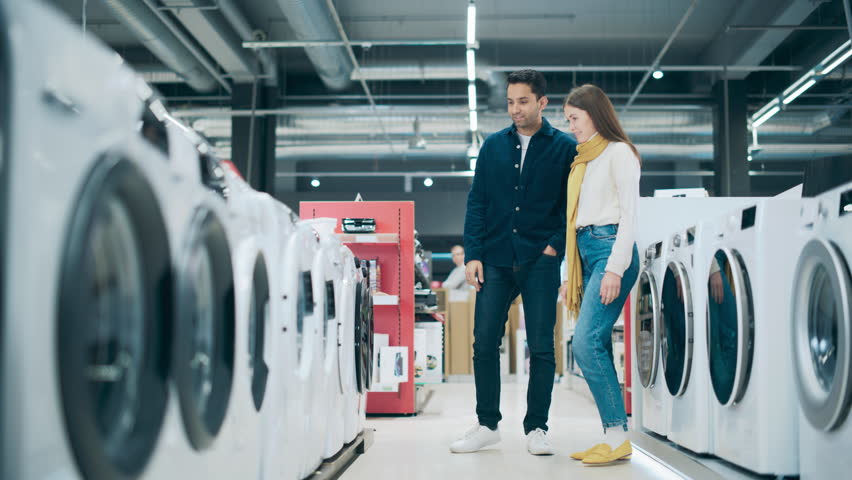 Young Multiethnic Couple Explores Home Electronics Store for Washing Machine Selection. Duo Shopping for Laundry Appliance. Customers Assess Modern Laundry Solutions in Contemporary Retail Shop
