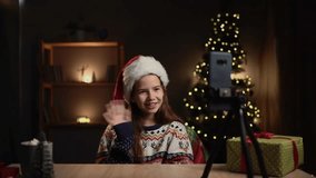 Teen girl sitting at table near Christmas tree and shooting her christmas video blog by smartphone on tripod. She wearing red santa claus hat.