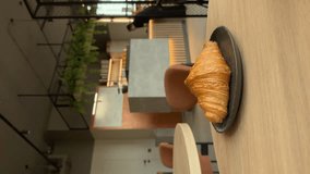 Eating fresh croissant with black coffee in a bakery. Dipping half of croissant into coffee, drops falling. People in bokeh, atmosphere of a coffee shop. Lifestyle vertical video.