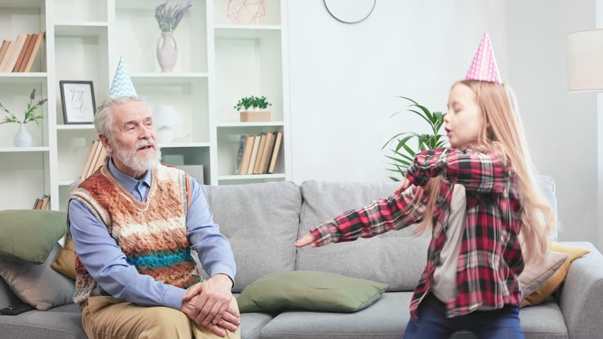Restless active girl dancing merrily in cozy living room. Casually dressed grey-bearded senior man catching positive vibe and having fun with grandchild. Concept of birthday celebration and happiness. Royalty-Free Stock Footage #1104387567
