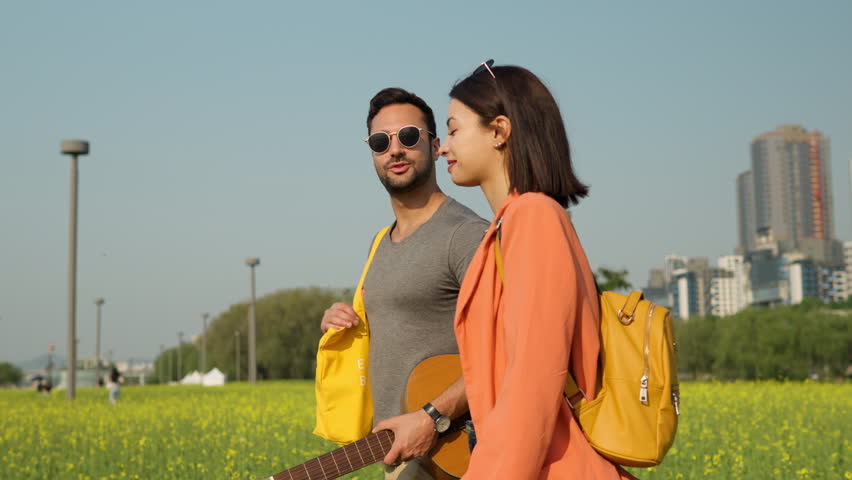 Happy Couple Man and Woman in Love Talking and Walking Throung Yellow Rapeseed Field in Bloom in Seorae Island by Han river Park in Seoul city - tracking side view in slow motion