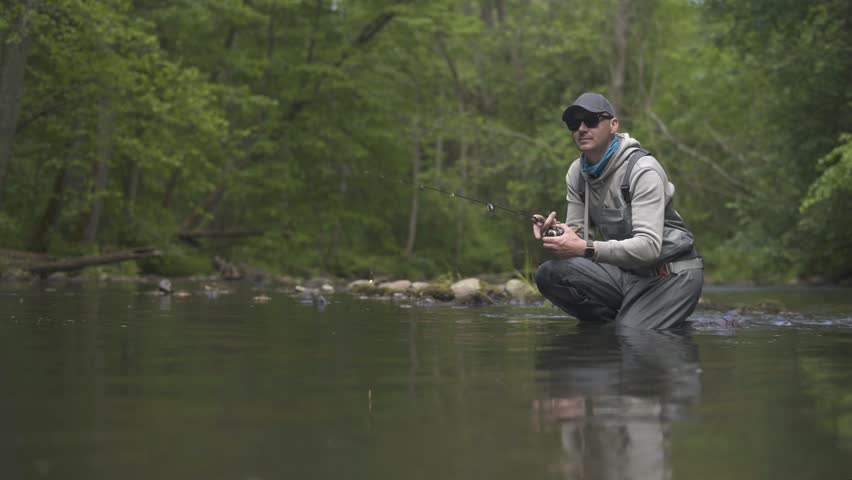 Fisherman catching brown trout on spinning tackle standing in river.