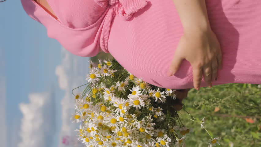 Vertical video. Close-up of young pregnant woman touching her belly in summer field, slow motion. Holding bouquet with daisies