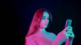 Happy beautiful girl with a smile  holds a smartphone and looks at the application in the studio on a colorful neon pink lights. Creative modern female portrait, 4k video footage