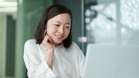 Smiling young adult asian woman using laptop while sitting at desk at workplace in modern office. A happy pretty female writes a text message, chats online, browsing dating site, communicates remotely - Powered by Shutterstock - Get 15% off with code: PIKWIZARD15