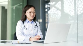 Asian female doctor talking on video call using laptop in hospital clinic. A smiling medical worker physician is having a conversation with a patient sitting at the desk at the workplace in the office