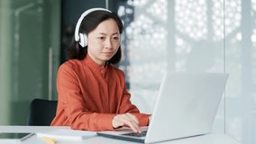 Asian woman in wireless headphones IT professional programmer working typing coding on laptop while sitting at workplace in modern office. Busy businesswoman developer analyzes financial digital data - Powered by Shutterstock - Get 15% off with code: PIKWIZARD15