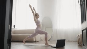 Girl in sports clothes performs yoga exercise using laptop to watch lesson in bright room. Slow motion