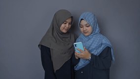 two asian women wearing hijab watching funny videos on smartphone and laughing in front of grey background in studio