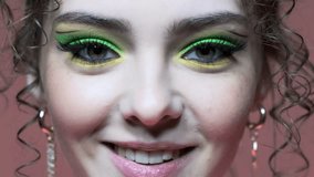 Сlose-up slow motion video portrait of young woman on pink background. Female with unusual green eyes shadows makeup, curly hair and earrings.