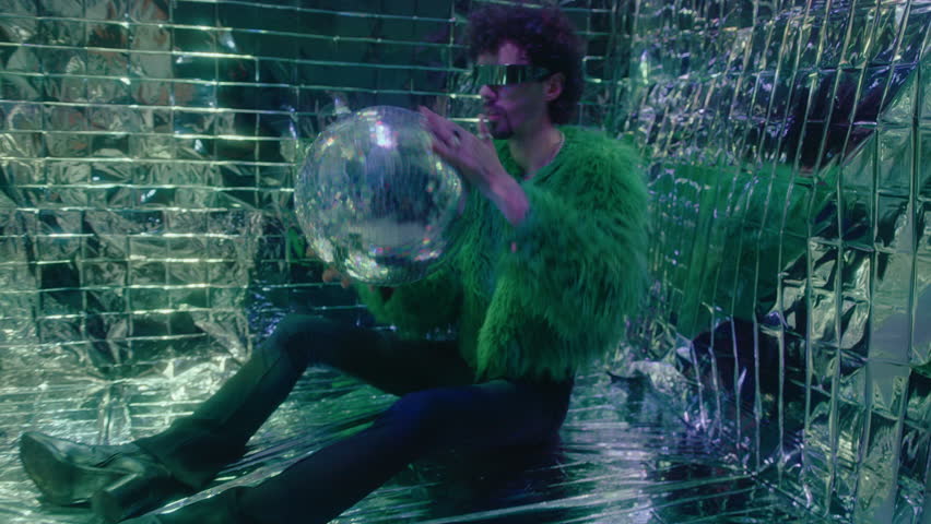 Trendy man wearing feather jacket, futuristic sunglasses and bell-bottoms sitting on floor in studio with colorful neon light and shiny background, posing on camera with disco ball. Zoom Shot