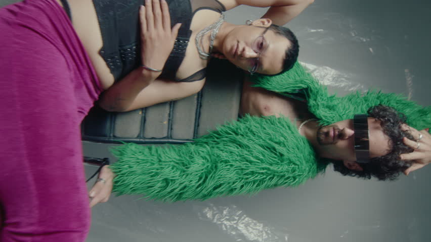 Young models wearing trendy clothing, eyewear and jewelry posing together for camera in light flashes against plastic wrap background, studio photoshoot for fashion magazine. Vertical clip