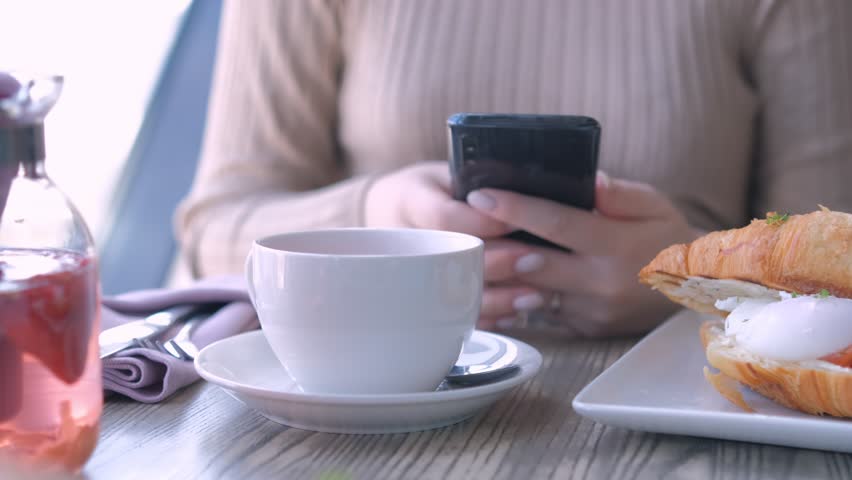 Unrecognizable businesswoman sitting at table in cafe cafeteria restaurant with a smartphone in hand prepare eat croissant with salmon and poached egg | Shutterstock HD Video #1104420921