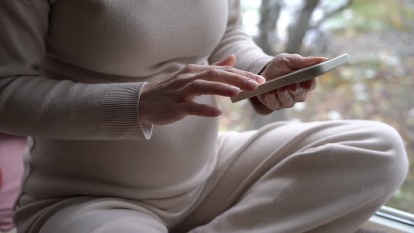 Unrecognizable pregnant woman scrolling apps on smartphone touchscreen sitting on windowsill. Happy relaxed Caucasian expectant messaging online surfing Internet in phone application | Shutterstock HD Video #1104423251