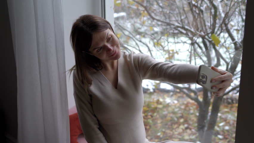 Cheerful pregnant young woman sitting on windowsill taking selfie with smartphone app. Smiling confident happy Caucasian expectant photographing gesturing enjoying pregnancy | Shutterstock HD Video #1104423253