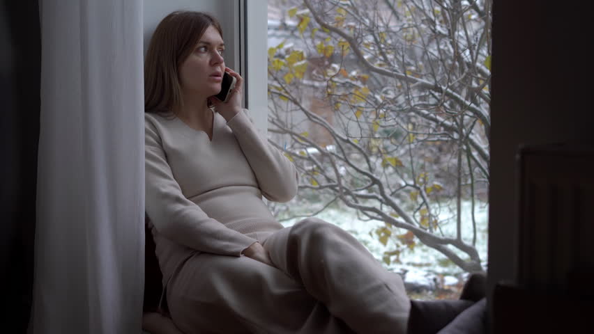 Young pregnant woman talking on phone sitting on windowsill indoors. Portrait of relaxed confident Caucasian expectant chatting on smartphone at home | Shutterstock HD Video #1104423259