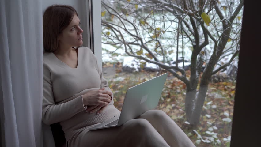 Young happy pregnant woman sitting on windowsill messaging online on laptop looking away. Portrait of satisfied relaxed Caucasian expectant enjoying leisure at home indoors | Shutterstock HD Video #1104423267