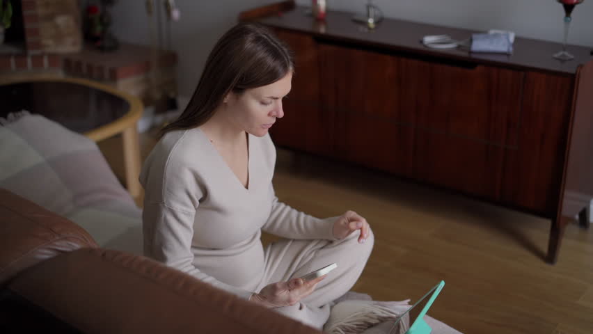 High angle view wide shot young pregnant woman scrolling digital tablet touchscreen dialing phone talking. Confident attractive Caucasian expectant calling sitting on couch in living room | Shutterstock HD Video #1104423299